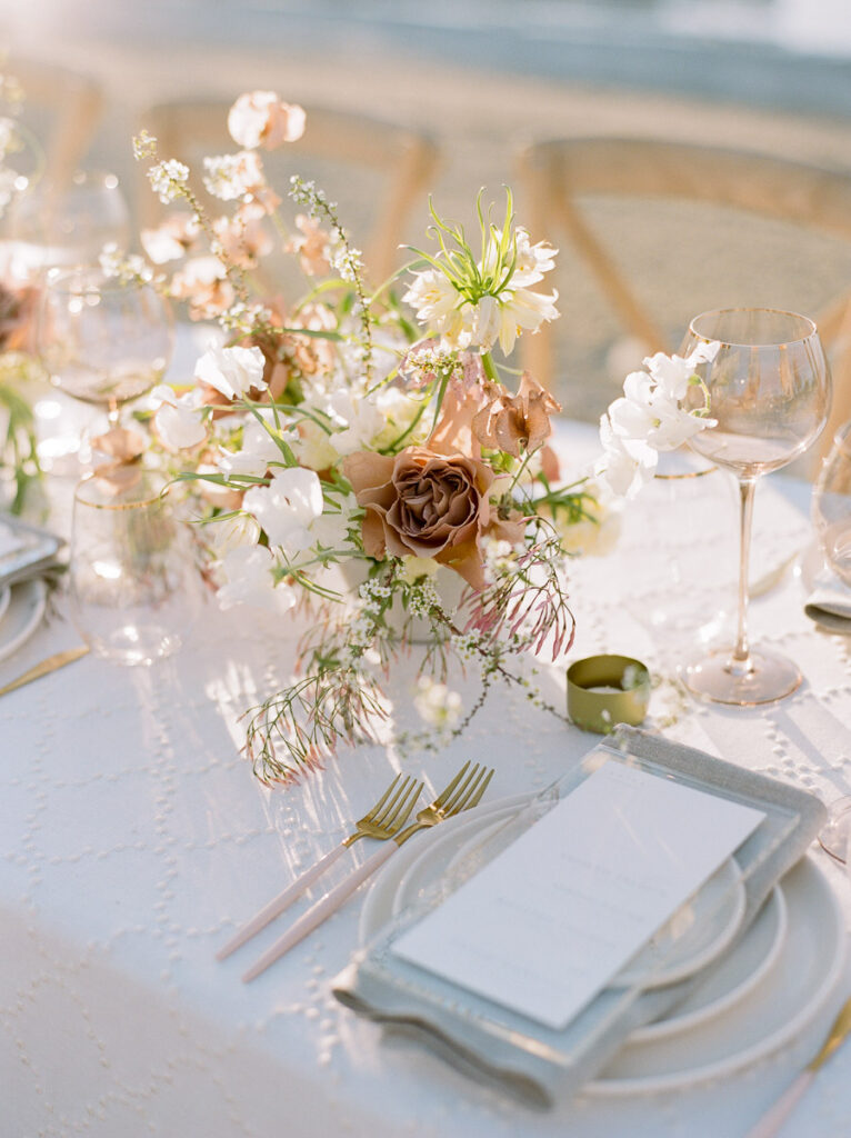 A wedding table layout featuring white textured pattern tablecloth, menus plates and napkins along with gold utensils and blush and white coloured florals.