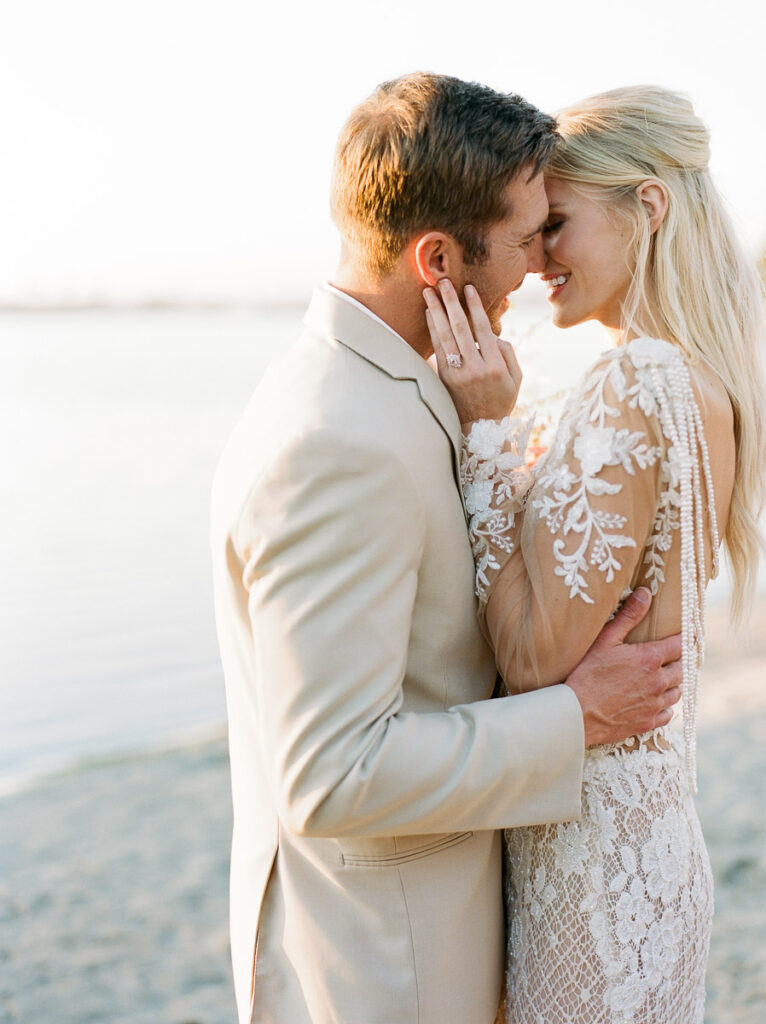 A bride and groom lean in for a kiss on a sandy San Diego beach. The bride is wearing a shear white lace wedding dress and a ring can be seen on her left hand. Her blonde hair is partially pulled back and hanging down her back loosely. The groom is in a cream coloured suit,
