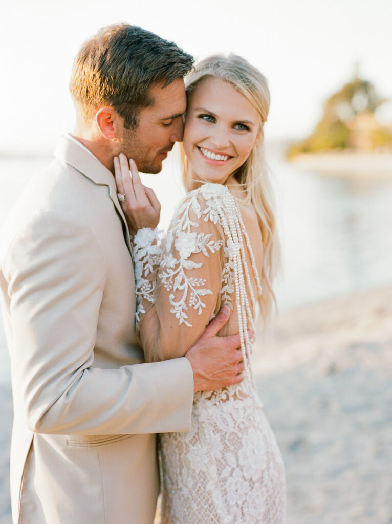 A bride and groom cuddle close on a  on a sandy San Diego beach. The groom looks like he is about to kiss the bride on the cheek as she smiles and looks away from him. The bride is wearing a shear white lace wedding dress and a ring can be seen on her left hand. Her blonde hair is partially pulled back and hanging down her back loosely. The groom is in a cream coloured suit,
