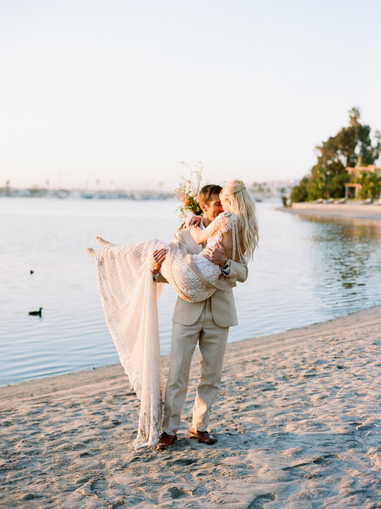 A groom in a cream suit pick up and craddels a bride in a shear white lace wedding dress, Her blonde hair is loose down her back. The pair are on a sandy San Diego beach,