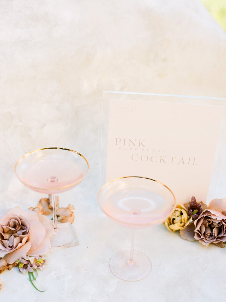 Two light pink drinks sit in a gold trimmed cocktail glass with a sign that says pink champagne cocktail. The glasses have blush coloured roses next to them
