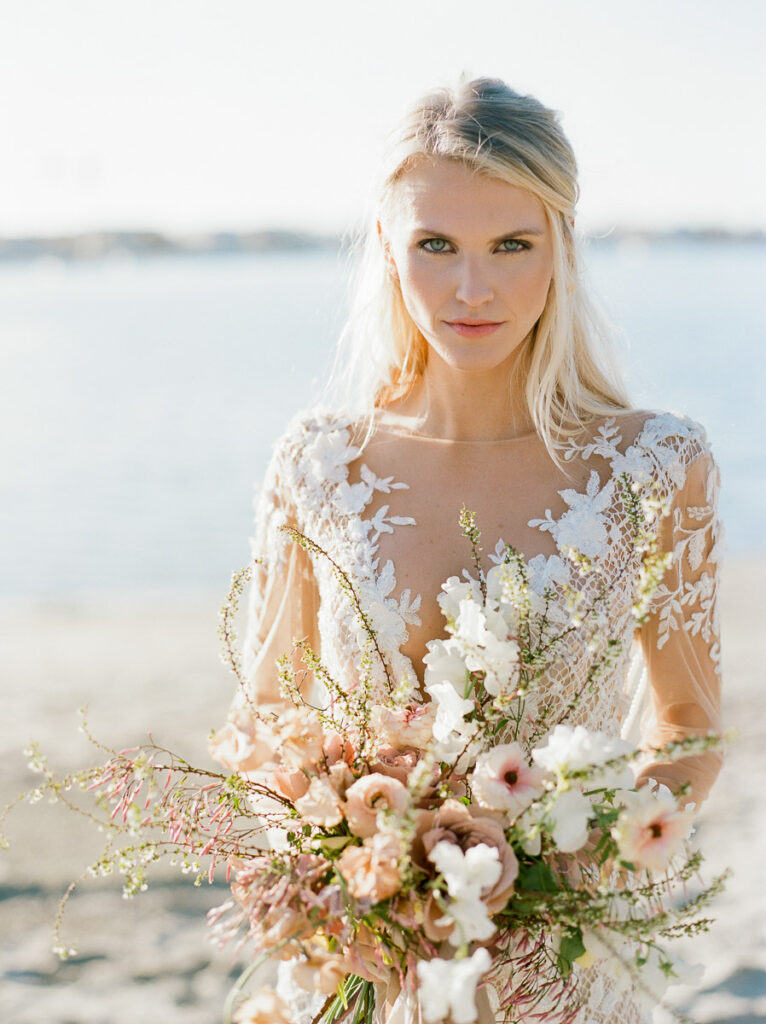 Blonde bride stands on a sandy San Diego beach with the water in the background. She is looking directly at the camera. She is wearing a detailed white lace wedding dress and is holding a large bridal bouquet with flowers in various shades of blush as well as white flowers and light coloured greenery.