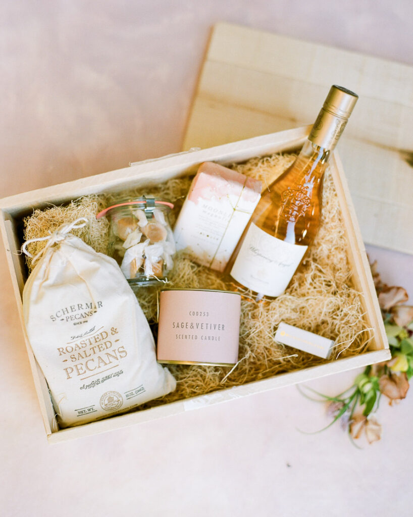 A welcome box filed with items such as a bottle of champaign, salted pecans, a scented candle, salt water toffee candies, and soap. All of the items are inside a wooden box and placed on top of straw filling.
