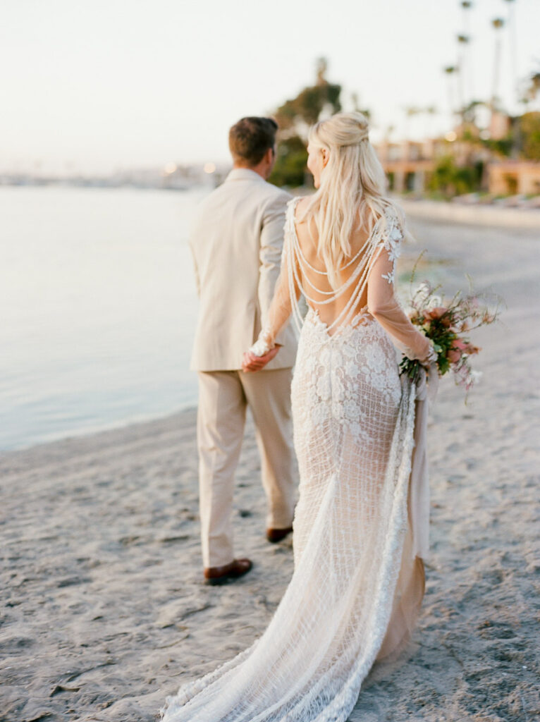 A bride and groom walk down a sandy San Diego beach with water in the background. The bride is in a shear white lace wedding dress and is holding a bouquet of flowers, the groom is in a cream suit. Both have their backs turned to the camera.