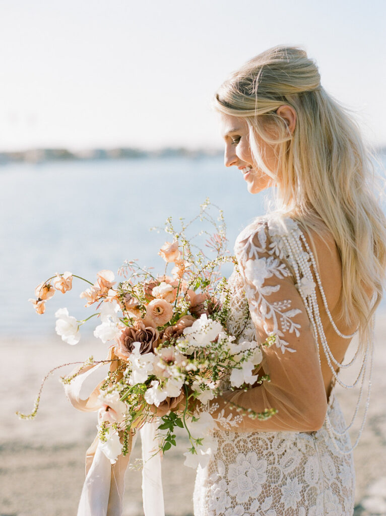 A bride is pictured from the waist up and she is angled away from the camera so all you can see is the side of her face. She is wearing white lace wedding Dress and is standing on a San Diego beach . She is holding a bouquet with light coloured greenery, both light and dark blush coloured flowers and white flowers. There are white and beige ribbons streaming down from the bouquet. The bride has her long. blonde hair partially tied up and the rest hanging loosely down her back.